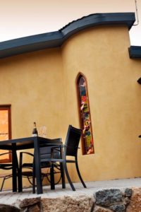 Paved verandah with table and chairs, the rounded outside wall of the chalet with a stained glass window